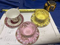 Photo showing three vintage tea cups and their matching saucers on a blue cloth, with measuring tape in frame indicating sizes.