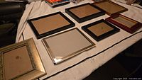 Seven picture frames laid out on white cloth, showing different sizes and colors, including black, silver, gold, brown, and burgundy. Frames are empty and facing upward with backing boards visible.