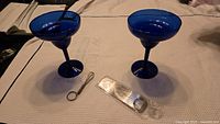 Two blue glass margarita glasses with a silver metal bottle opener and a keychain whisk on a white textured cloth.