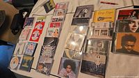 Overview photo showing a collection of approximately 34 CDs laid out on a flat surface, including different genres with intact jewel cases and minor signs of wear.