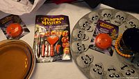 Photo showing brown Pyrex pie plate, pumpkin carving tool set in packaging, LED strobe light, black Halloween bucket with orange bead handle, and skull decorative platter.