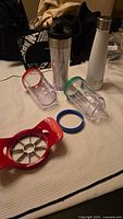 Photo showing white stainless steel flask, two plastic fruit infuser parts with colored rims, and red apple slicer on fabric surface