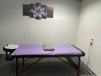 Side view of the mobile massage table with purple disposable sheet and gray towel on top, showing the adjustable armrest side attachment and face cradle.