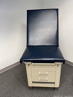 Full front view of one medical exam chair showing dark blue padded seat and backrest on beige cabinet with drawers.