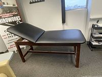 Photo of one massage table featuring dark wood frame and black padded adjustable backrest angled upwards.