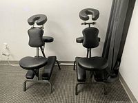 Two black mobile massage chairs set up with adjustable padded headrests, armrests, chest support, and cushioned seats on metal frames.