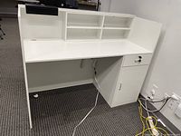 Front view of white reception desk showing work surface, hutch shelving, lockable drawer, and door cabinet below.
