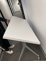 White adjustable table with metal tripod base, smooth white rectangular top, held by person for scale.