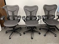 Front view of three black mesh office chairs with ergonomic backrests and armrests spaced side by side on carpet.