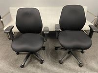 Two black ergonomic office chairs with cloth upholstery and adjustable armrests, positioned side by side on a carpeted floor in front of a white couch.