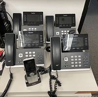 Five Yealink phones displayed on a table including four wired desk phones with LCD screens and one cordless phone on a charging dock, visible cords and network switch nearby.