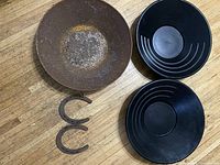 All five items including a large rusted metal gold pan, two smaller black plastic gold pans, and two rusted metal horseshoes laid out on a wooden floor.