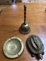 Photo showing the three brass items on a wooden surface: hand bell, ashtray, and turtle figurine.