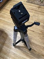 Black Ambico tripod showing full legs with protective plastic wrap and head mounted on floor background.