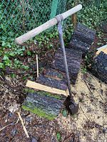 Photo showing the full antique hand auger leaning against wood pieces and greenery, with visible wooden T handle and long metal shaft ending in square metal digging blade