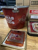 Red Budweiser beer bucket with Gordon's Dry Gin mirror sign in front on wooden table, cardboard boxes in background