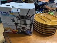 Home Outfitters 10-piece stainless steel meat fondue set box and 6 yellow Staub divided plates stacked