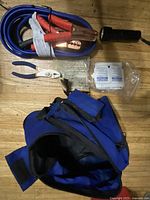 Top down photo of emergency tools and supplies spread around blue nylon bag on a wooden floor, showing jumper cables, flashlight, pliers, and gauze pads.