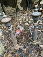 Pair of cast iron andirons with finials placed outdoors on dirt and rocks showing weathered condition.