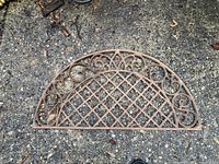 Top-down photo of half-round cast iron door mat, showing lattice and scrollwork pattern on textured concrete ground.