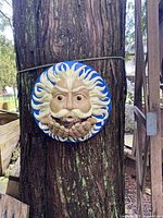 Front view of the ceramic sun plaque hanging on a tree, showing the sun face and hands cupped at the front.