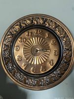 Front view of round copper wall clock showing raised floral pattern around edge and sunburst patterned clock face.