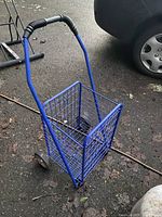 Lightweight blue metal grocery cart with wire mesh basket and black foam handle grip, showing the front and side view with four caster wheels on asphalt floor.