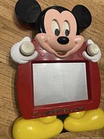 Front view of the Mickey Mouse Etch A Scratch drawing toy showing Mickey's face, ears, hands holding knobs, and feet with the screen in the center