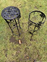 Pair of black metal plant stands on grass, one with solid circular top and floral cutout, other with open top and leaf accents.