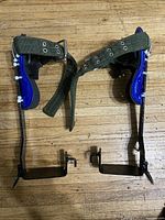 Top view of new tree climbing spurs showing blue padded foot support, green adjustable straps, and black metal frame with spikes.