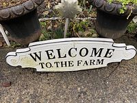 Photo of a white wooden welcome sign with black lettering 'WELCOME TO THE FARM' placed between plant pots outdoors on textured ground.