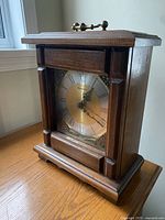 Front angled view of wooden mantel clock showing brass-colored clock face with Roman numerals, black hands, glass front, and decorative brass handle on top.