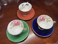Three Aynsley porcelain cups and saucers on a wooden surface: two matching with white cups featuring a pink rose and one with a coral cup with floral interior pattern.
