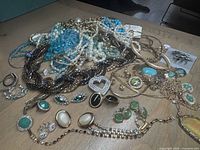 Top-down view of a mixed pile of costume jewellery on a wooden table, showing necklaces, earrings, and bracelets with pearls, turquoise, green stones, rhinestones, and gold-tone chains.