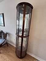 Full view of tall curved glass curio cabinet with wooden frame and glass shelves inside, showing the cabinet standing against a wall in an empty room.