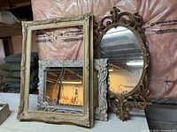 Three items displayed against a wall: large ornate gold-tone frame, smaller white ornate framed rectangular mirror, and large gold-tone oval framed mirror with intricate scroll design.