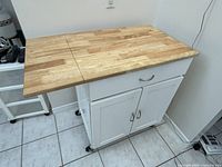Front-right angled view of mobile kitchen island showing butcher block wood top, drawer, cabinet doors, and caster wheels.