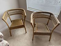 Two wicker chairs positioned side by side in a corner near a window. Both feature light brown wicker with curved backs and armrests.