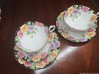 Two sets of Paragon teacups with matching saucers and one additional plate, all decorated with colorful chrysanthemum flowers on white china with black and gold trim, arranged on wood surface.