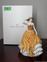 Royal Doulton Pretty Ladies Patricia porcelain figurine displayed with the original white box marked with Royal Doulton logo and 'England 1815'. Figurine is of a woman in an orange-yellow dress with a hat, holding the brim with one hand and lifting the dress with the other.