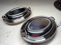 Two large silverplate covered serving dishes with handles and decorative edges.