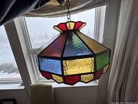 Stained glass hanging light fixture shown being held by hand near window with snowy outdoor view. Features hexagonal shape with multi-colored glass panels in red, blue, yellow, green, and pink.