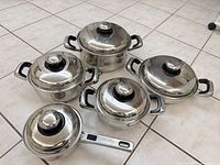Top-down angled view of five pieces of shiny Carrera stainless steel cookware including three pots, one saucepan and an oval pot, all with lids featuring black knobs.