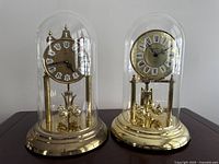 Two dome-covered anniversary clocks displayed side-by-side, showing their overall size and condition.