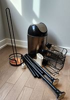 Bathroom accessories arranged with natural lighting: black metal toilet paper holder, metallic garbage can with dome lid, spare toilet paper rolls, toothbrush holders, and caddy components on floor