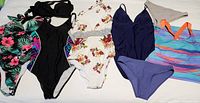 Seven items of women's beachwear displayed flat: Merrell multicolor sleeveless dress, two pairs of Romwe bikini bottoms, and four swimsuits including black George one-piece, white halter, blue lace-up back, floral halter one-piece, plus a black halter bikini top.