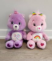Two Care Bear plush toys side by side on wooden floor against white wall. Purple Share Bear on left with crossed lollipop belly badge. Pink Cheer Bear on right with rainbow belly badge and rainbow bow.