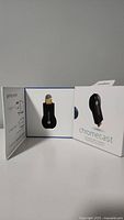 Black Chromecast device visible inside open original packaging box, device is securely inset in white plastic holder, box shows illustrations of device use and setup on inner flap.