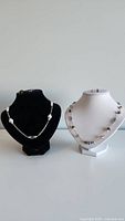 Two handmade beaded silver-tone necklaces displayed on black and white bust stands, showing bead and chain details