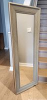 Full view of the grey framed rectangular mirror standing on floor beside stairway, showing frame color and size